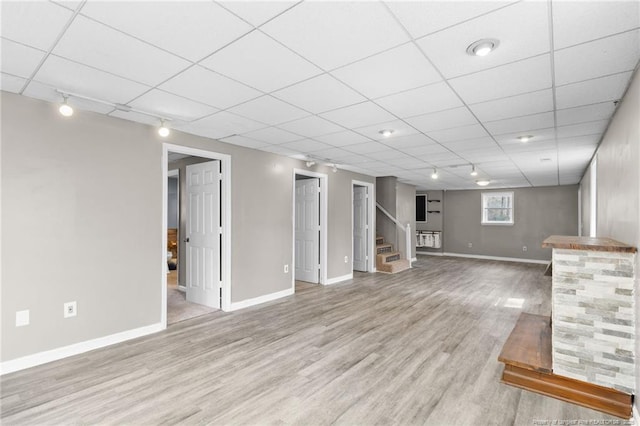 basement featuring light wood-style floors, a paneled ceiling, stairway, and baseboards