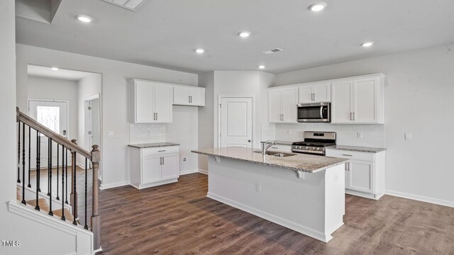 kitchen with dark wood-type flooring, white cabinetry, stainless steel appliances, light stone counters, and a center island with sink