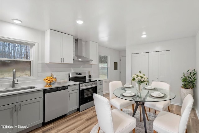 kitchen featuring gray cabinetry, light wood-type flooring, stainless steel appliances, wall chimney range hood, and sink