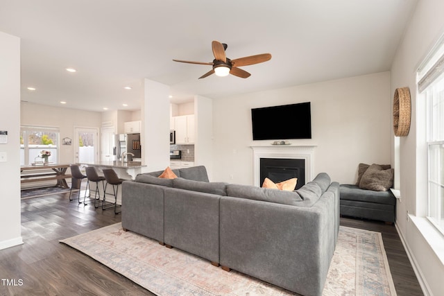 living room featuring dark wood-type flooring, a glass covered fireplace, recessed lighting, baseboards, and ceiling fan