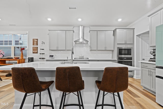 kitchen featuring gray cabinetry, oven, ornamental molding, a kitchen island with sink, and wall chimney exhaust hood