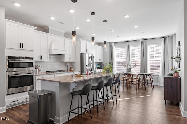 kitchen with white cabinets, hanging light fixtures, custom exhaust hood, stainless steel appliances, and a center island with sink