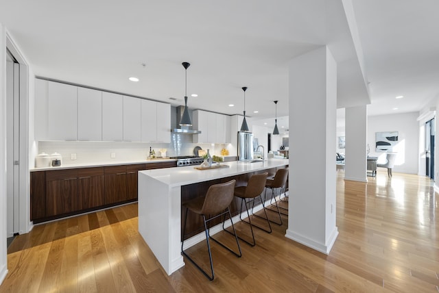 kitchen featuring wall chimney range hood, a kitchen island with sink, dark brown cabinetry, white cabinets, and decorative light fixtures