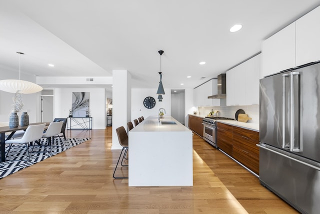 kitchen featuring wall chimney range hood, high end appliances, pendant lighting, and a center island with sink