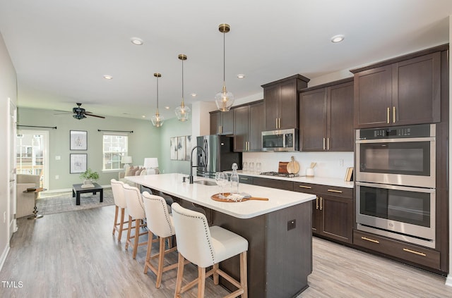 kitchen with appliances with stainless steel finishes, a center island with sink, light countertops, hanging light fixtures, and dark brown cabinetry