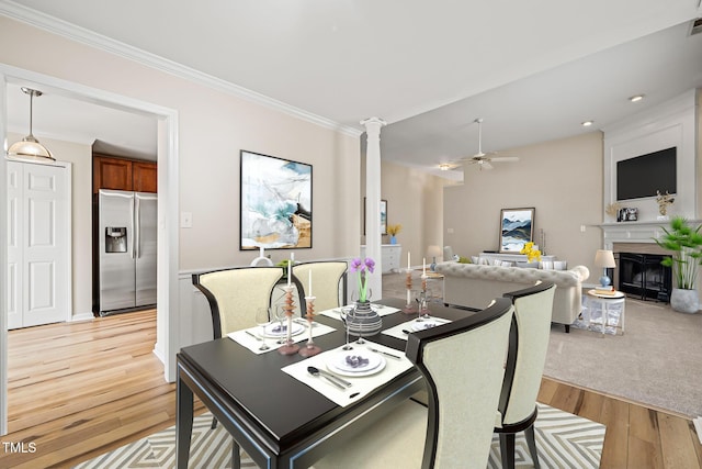 dining area featuring decorative columns, a fireplace with flush hearth, ceiling fan, crown molding, and light wood-style floors