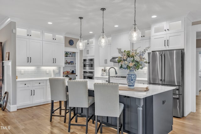 kitchen with stainless steel appliances, white cabinetry, an island with sink, glass insert cabinets, and pendant lighting