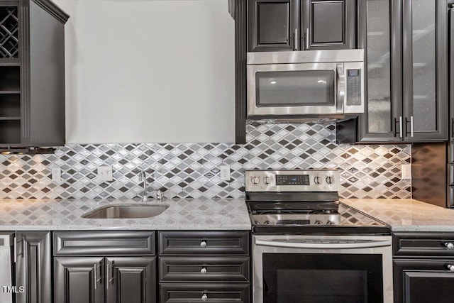 kitchen featuring stainless steel appliances, a sink, backsplash, light stone countertops, and glass insert cabinets