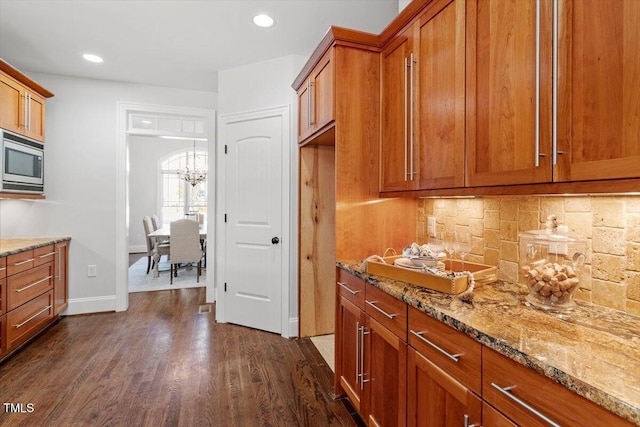 kitchen with decorative backsplash, stainless steel microwave, brown cabinets, dark wood-style flooring, and light stone countertops