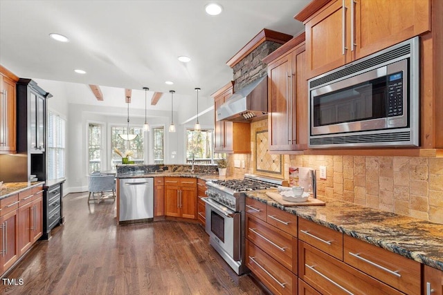 kitchen with pendant lighting, appliances with stainless steel finishes, brown cabinetry, wall chimney range hood, and dark stone counters