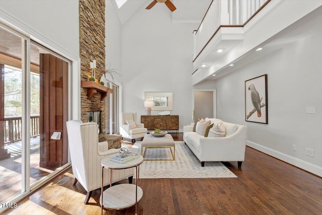 living room featuring baseboards, a ceiling fan, dark wood-type flooring, a high ceiling, and a stone fireplace