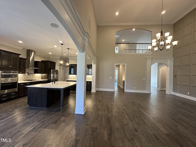 kitchen with pendant lighting, light countertops, appliances with stainless steel finishes, wall chimney range hood, and an island with sink