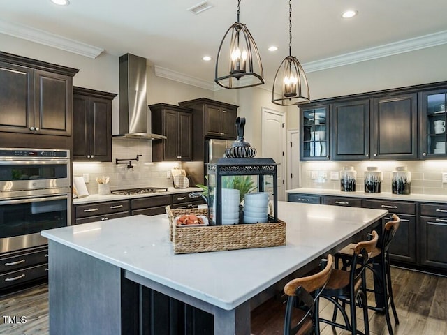 kitchen featuring an island with sink, wall chimney exhaust hood, appliances with stainless steel finishes, and glass insert cabinets