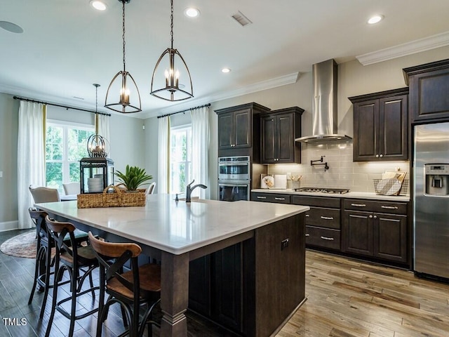 kitchen with light countertops, wall chimney range hood, hanging light fixtures, appliances with stainless steel finishes, and a large island with sink