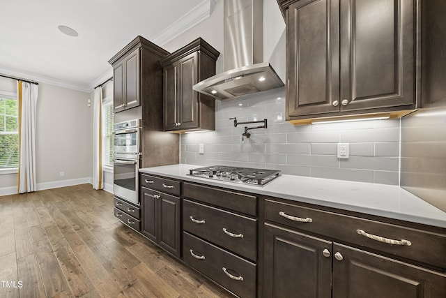 kitchen with wall chimney exhaust hood, crown molding, dark brown cabinetry, baseboards, and appliances with stainless steel finishes