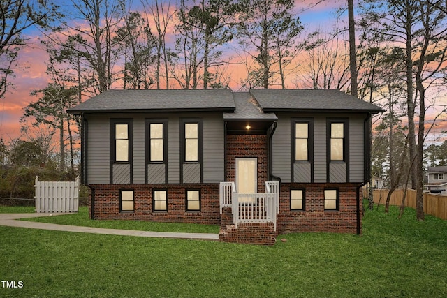 split foyer home featuring a yard, brick siding, roof with shingles, and fence