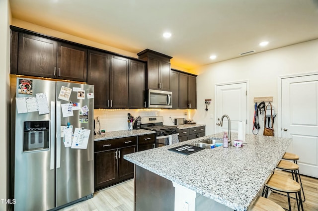 kitchen with appliances with stainless steel finishes, sink, light stone countertops, and an island with sink