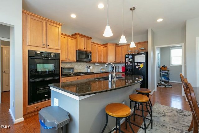 kitchen with a center island with sink, tasteful backsplash, a sink, wood finished floors, and black appliances