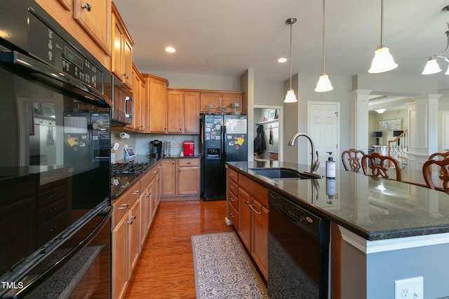 kitchen with a sink, brown cabinetry, black appliances, an island with sink, and ornate columns