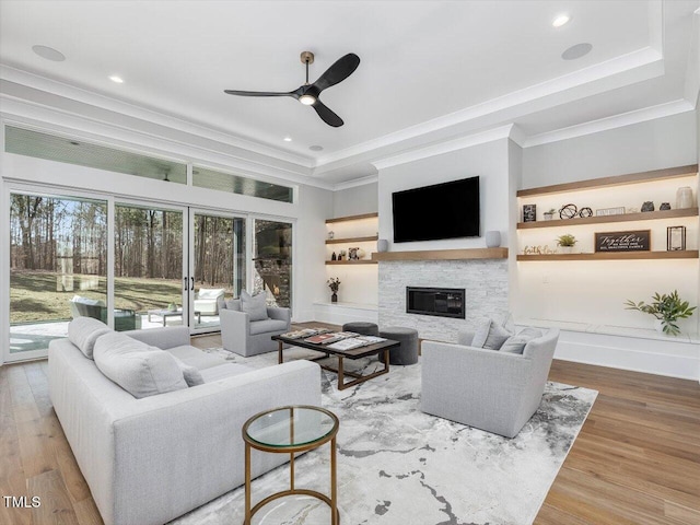 living room with a tray ceiling, crown molding, ceiling fan, a stone fireplace, and wood finished floors