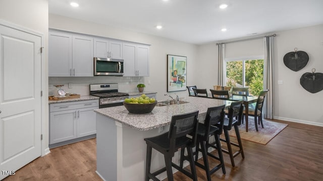 kitchen featuring a breakfast bar, hardwood / wood-style floors, stainless steel appliances, light stone counters, and an island with sink