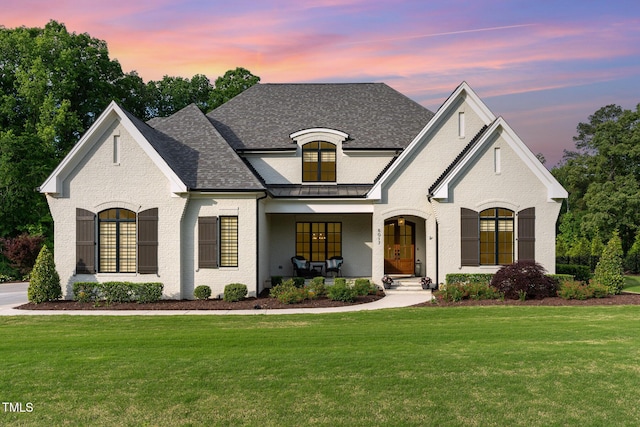 french country style house with covered porch, brick siding, a shingled roof, and a front lawn