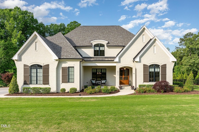 french country home with covered porch, brick siding, a front yard, and roof with shingles