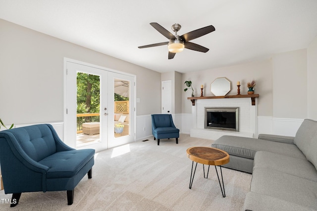 living room featuring light colored carpet, a fireplace, french doors, a wainscoted wall, and a ceiling fan