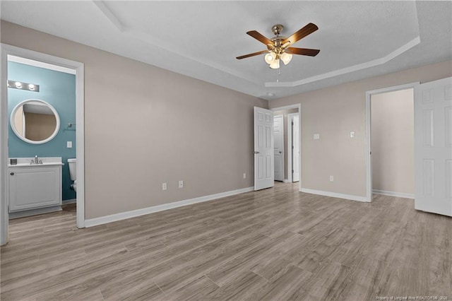 unfurnished bedroom with sink, ensuite bath, light wood-type flooring, a tray ceiling, and ceiling fan