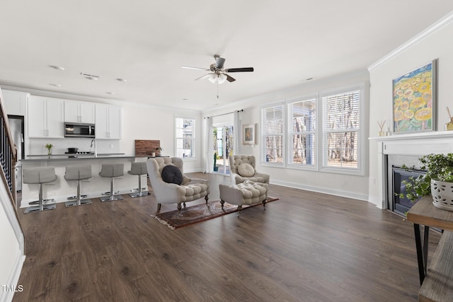 living area featuring dark wood-style flooring, crown molding, ceiling fan, a tile fireplace, and baseboards