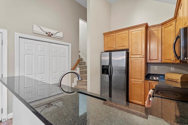 kitchen with black microwave, a sink, stainless steel refrigerator with ice dispenser, brown cabinetry, and dark stone countertops