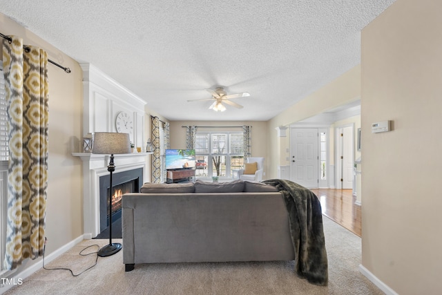 living room featuring ceiling fan, a textured ceiling, light colored carpet, baseboards, and a lit fireplace