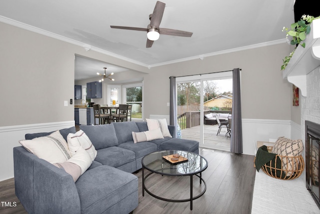 living area with a brick fireplace, crown molding, dark wood-type flooring, a wainscoted wall, and ceiling fan with notable chandelier