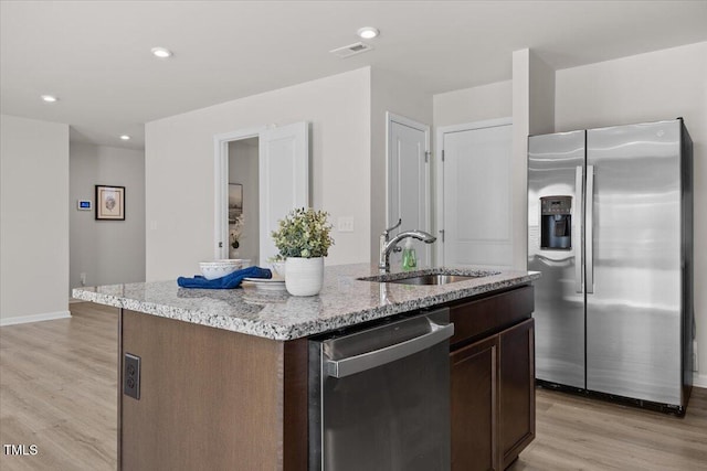 kitchen with light wood-style flooring, stainless steel appliances, a sink, dark brown cabinets, and a center island with sink