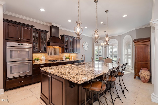 kitchen featuring an island with sink, custom range hood, a sink, stainless steel appliances, and decorative backsplash