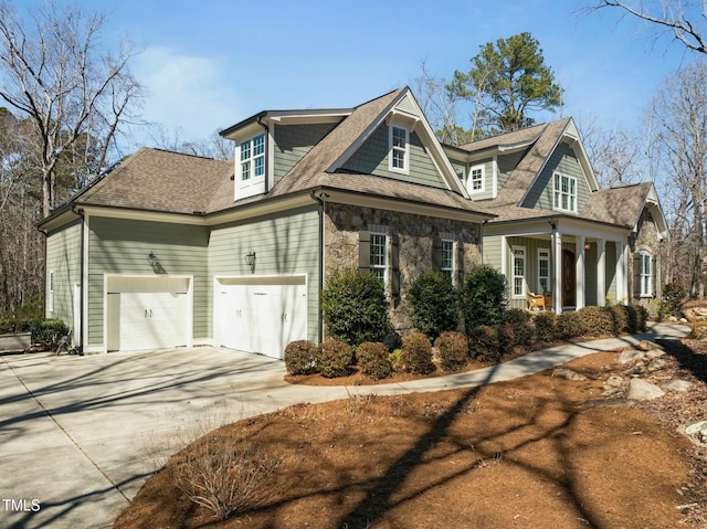 shingle-style home featuring a porch, an attached garage, stone siding, concrete driveway, and roof with shingles