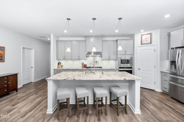 kitchen with appliances with stainless steel finishes, hanging light fixtures, light stone counters, and a center island with sink