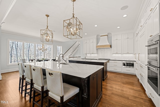 kitchen with a sink, an island with sink, an inviting chandelier, and premium range hood