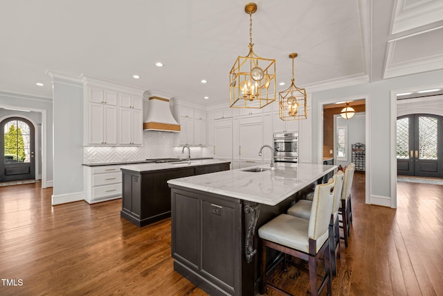 kitchen with premium range hood, an island with sink, a sink, a notable chandelier, and backsplash