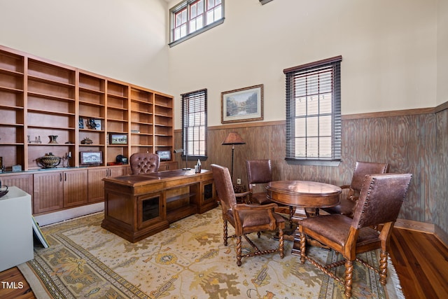 office area with light wood finished floors, a towering ceiling, wood walls, and wainscoting