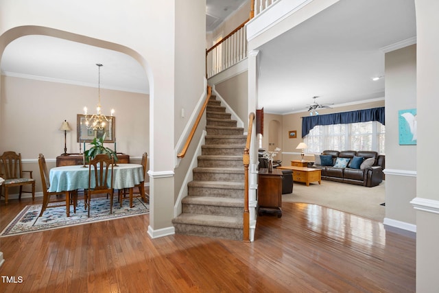 staircase with arched walkways, ornamental molding, wood finished floors, baseboards, and ceiling fan with notable chandelier