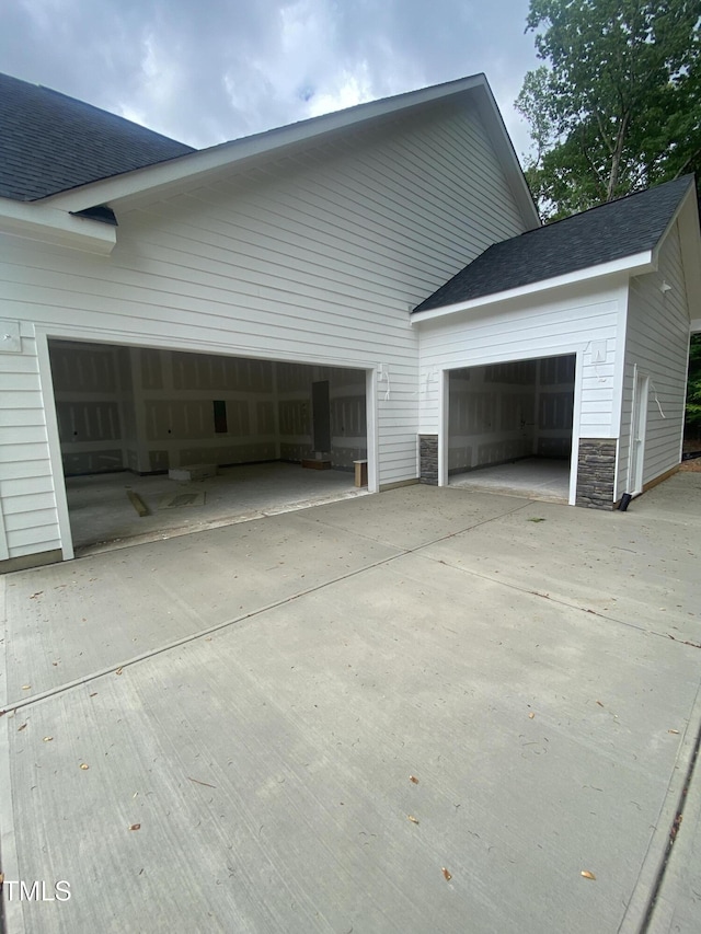 view of side of property with stone siding, an attached garage, driveway, and roof with shingles