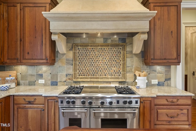 kitchen featuring premium range hood, range with two ovens, light stone counters, decorative backsplash, and brown cabinets