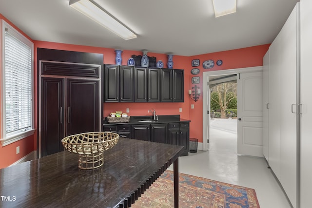 kitchen with a sink, dark countertops, finished concrete floors, baseboards, and paneled built in refrigerator