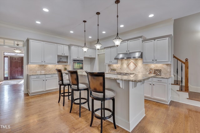 kitchen with hanging light fixtures, appliances with stainless steel finishes, a kitchen island, light stone countertops, and under cabinet range hood