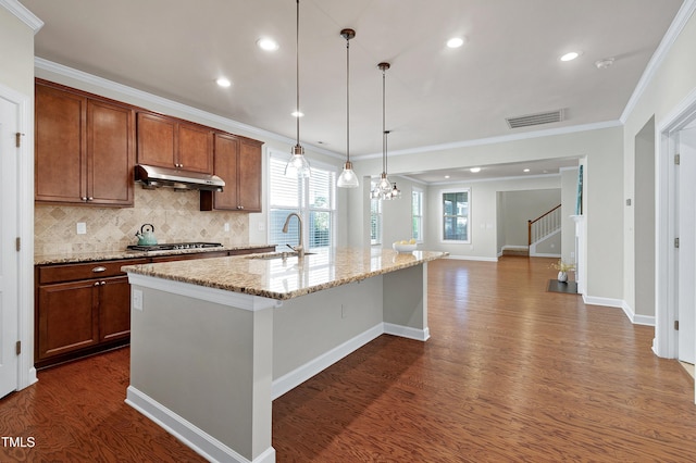 kitchen with a center island with sink, hanging light fixtures, sink, light stone counters, and dark wood-type flooring