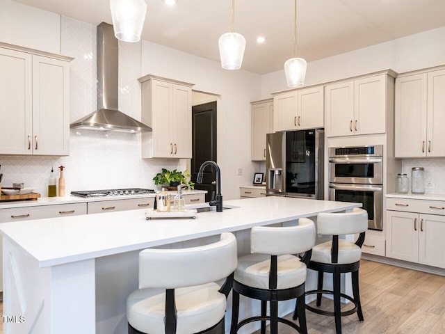 kitchen featuring hanging light fixtures, appliances with stainless steel finishes, light countertops, wall chimney exhaust hood, and a center island with sink