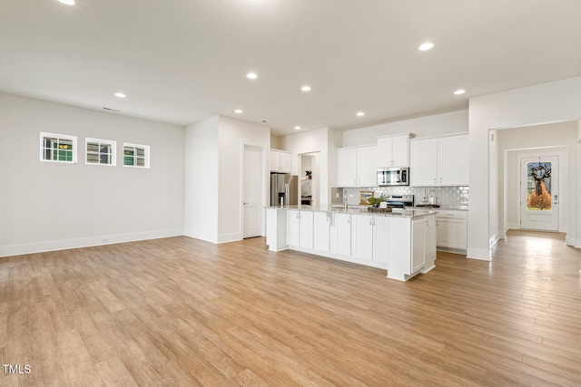 kitchen featuring white cabinets, appliances with stainless steel finishes, light wood finished floors, a center island with sink, and tasteful backsplash