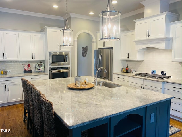 kitchen with a kitchen island with sink, stainless steel appliances, a sink, white cabinetry, and hanging light fixtures