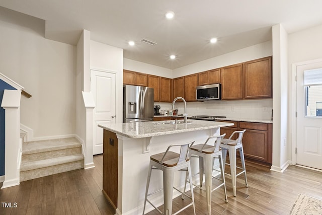 kitchen featuring dark wood-style floors, stainless steel appliances, visible vents, brown cabinetry, and an island with sink